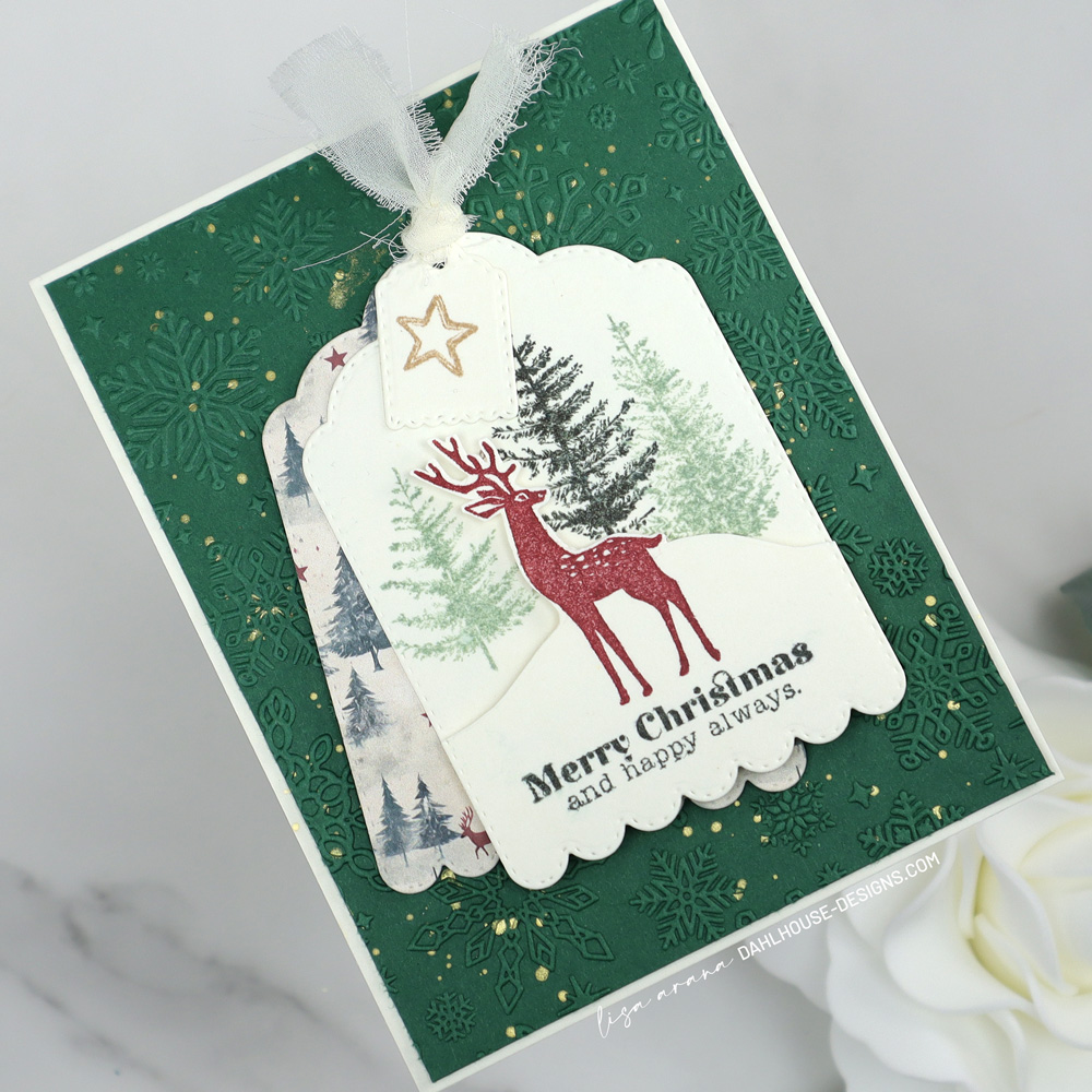 Layered Christmas card featuring a scalloped tag with stamped trees, a red deer, and a Merry Christmas sentiment, layered over patterned paper and set on an embossed green snowflake background with gold splatter. designed by Lisa Arana dahlhouse designs dot com