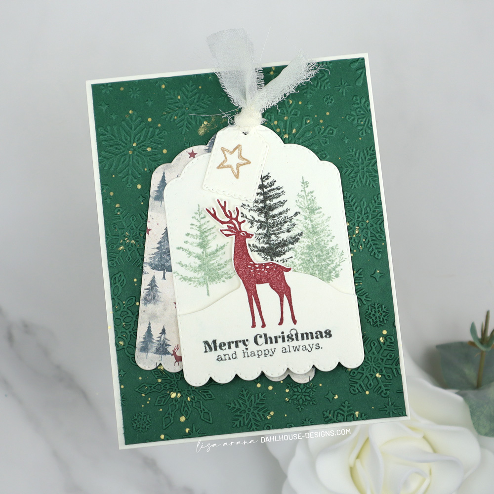 Layered Christmas card featuring a scalloped tag with stamped trees, a red deer, and a Merry Christmas sentiment, layered over patterned paper and set on an embossed green snowflake background with gold splatter. designed by Lisa Arana dahlhouse designs dot com