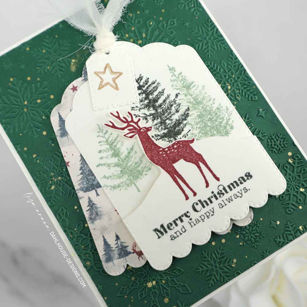 Layered Christmas card featuring a scalloped tag with stamped trees, a red deer, and a Merry Christmas sentiment, layered over patterned paper and set on an embossed green snowflake background with gold splatter. designed by Lisa Arana dahlhouse designs dot com